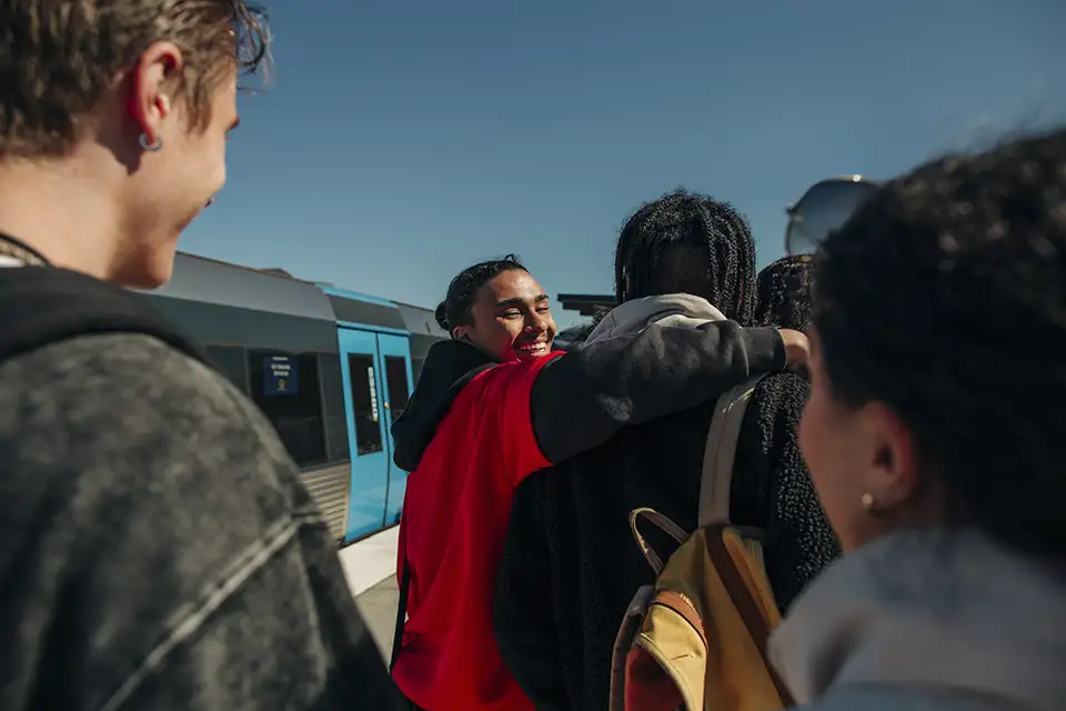 Ungdomar med utländsk bakgrund håller om varandra och ler på en tågstation.
