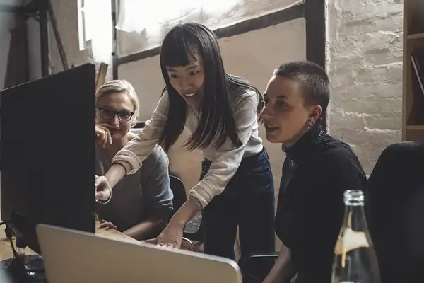 Tre kvinnor pratar framför en datorskärm på ett kontor.