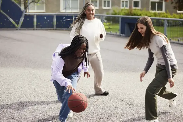 Tre tonårsflickor spelar basket på skolgård.
